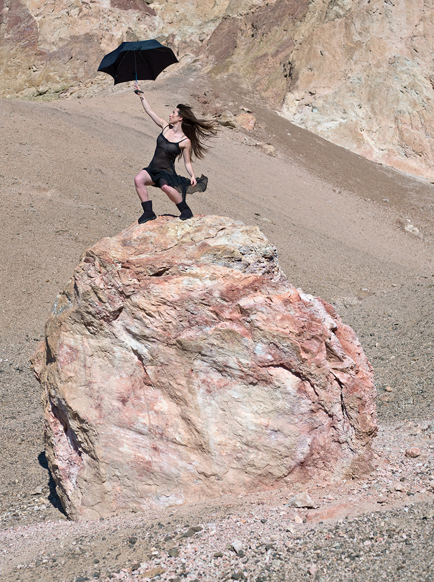 Woman Standing on the top of a large rock in Death Valley holding an Umbrella and being blown by the wind.
